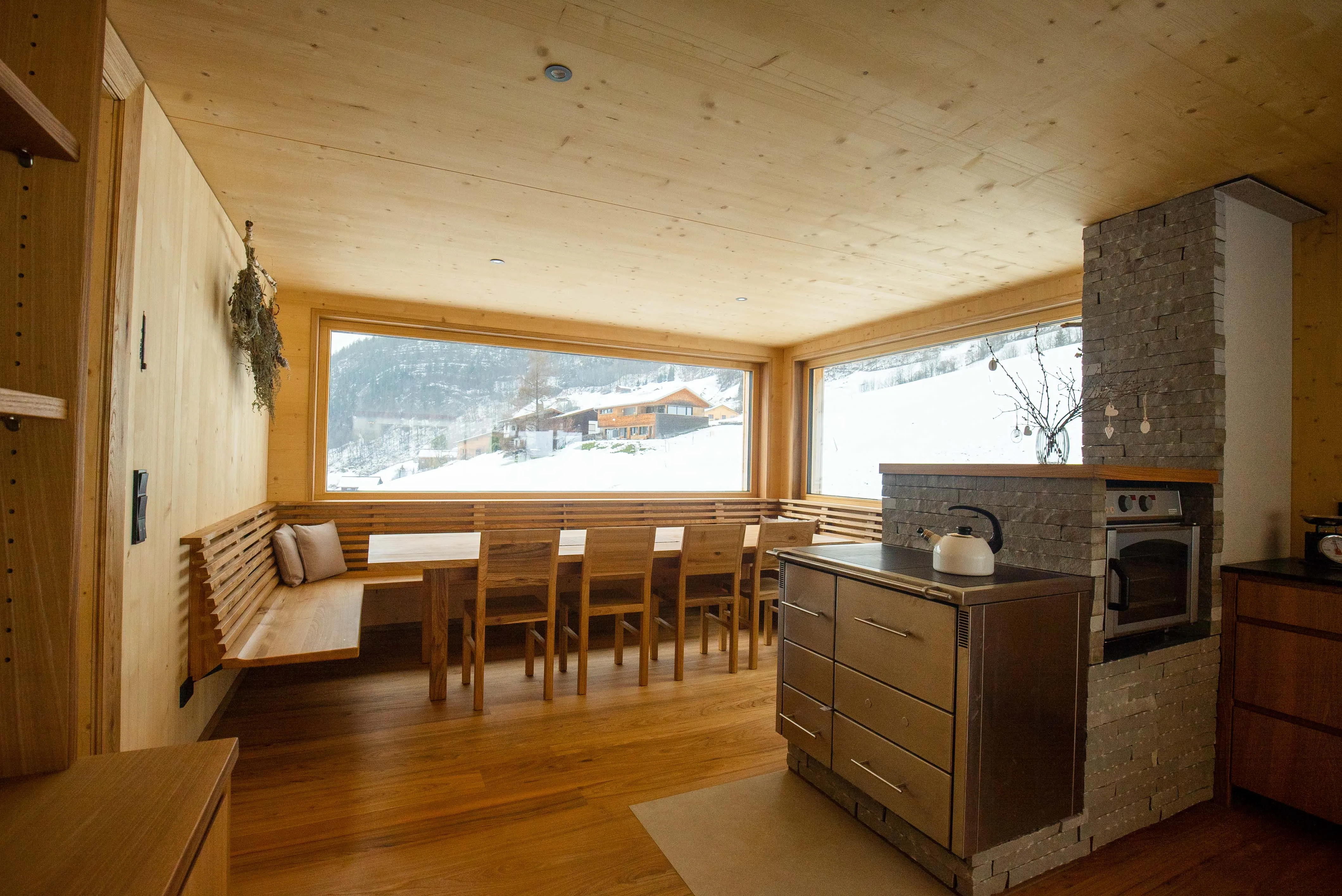 Heller Seminarraum aus Holz mit langem Tisch, Eckbank, Holzofen und Blick auf die verschneiten Berge